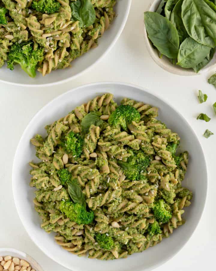 two bowls of vegan broccoli pesto pasta surrounded by a bowl of basil and a bowl of toasted pine nuts