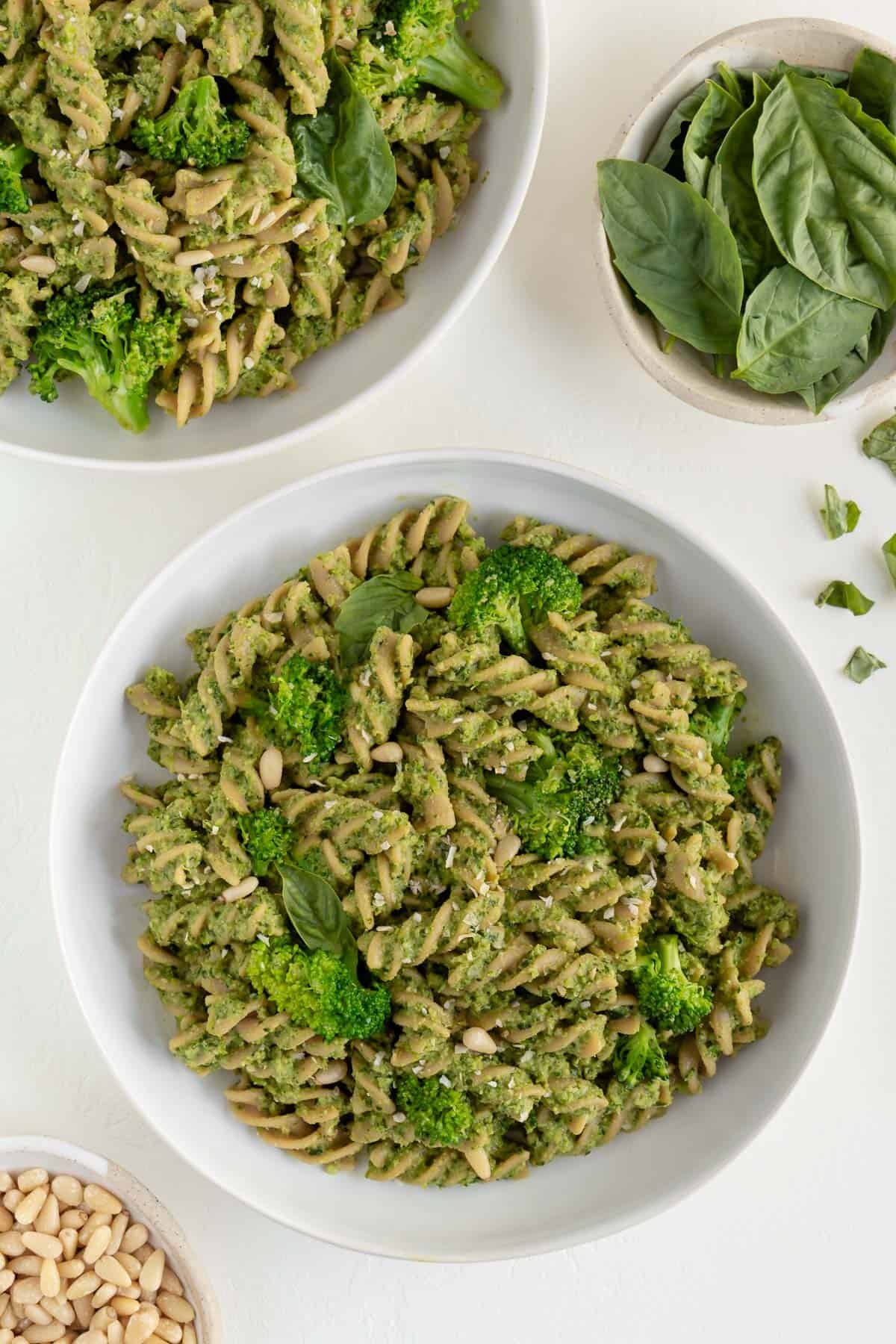 two bowls of vegan broccoli pesto pasta surrounded by a bowl of basil and a bowl of toasted pine nuts