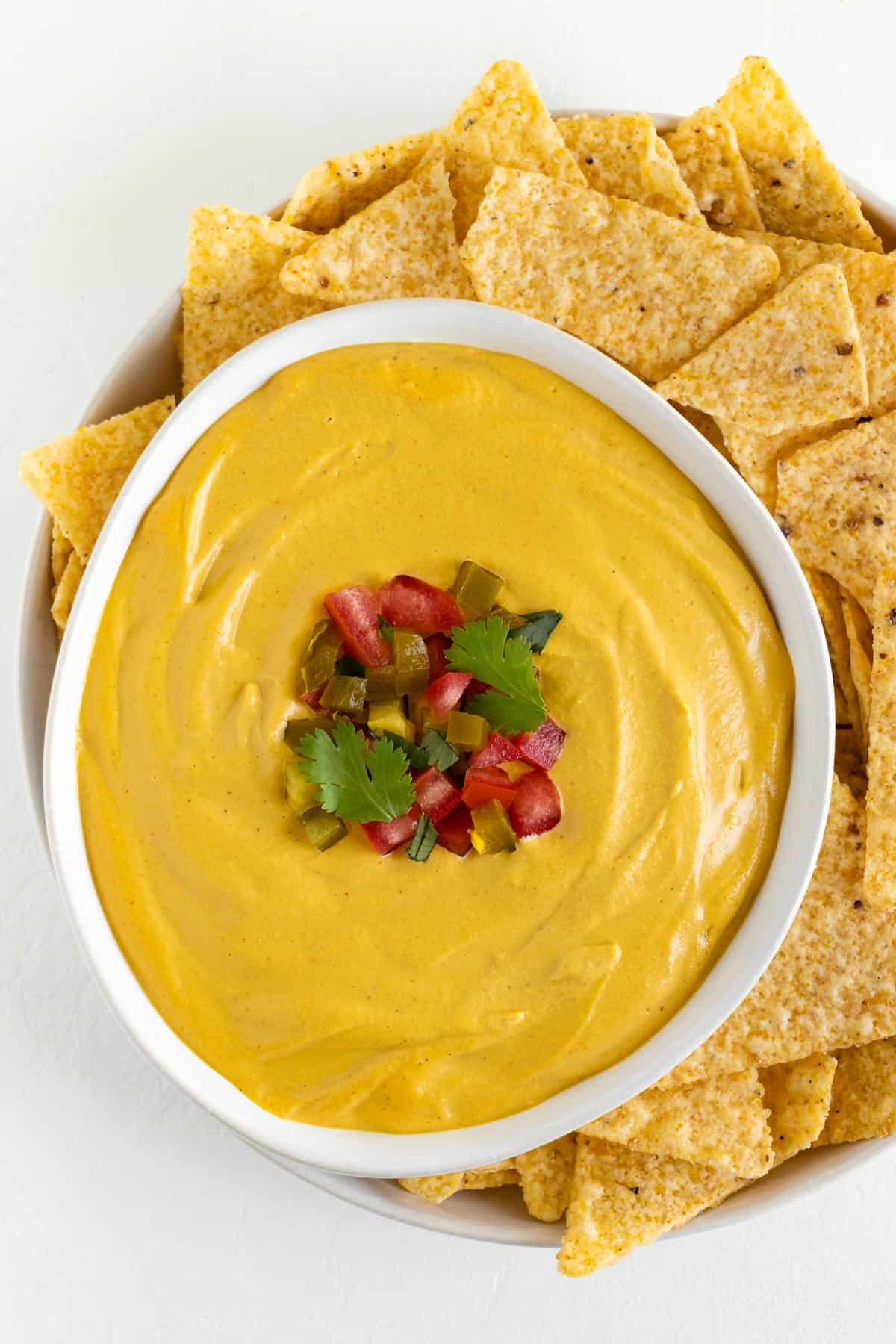 a bowl of vegan nacho cheese garnished with diced jalapeño, tomato, and cilantro beside corn tortilla chips