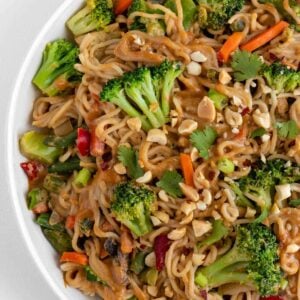 vegan peanut noodles in a white bowl with broccoli, carrots, peas, mushrooms, and bell peppers