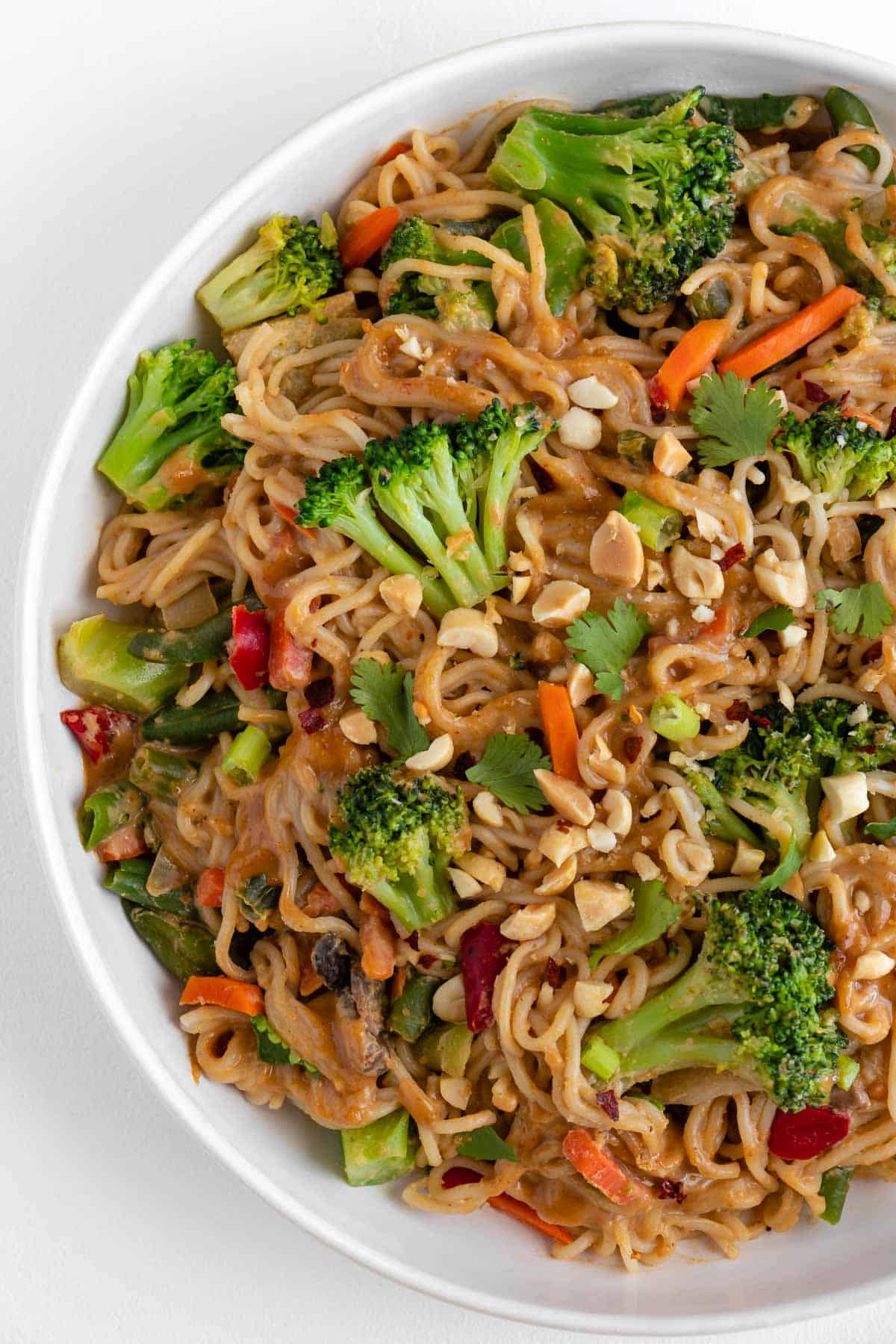 vegan peanut noodles in a white bowl with broccoli, carrots, peas, mushrooms, and bell peppers