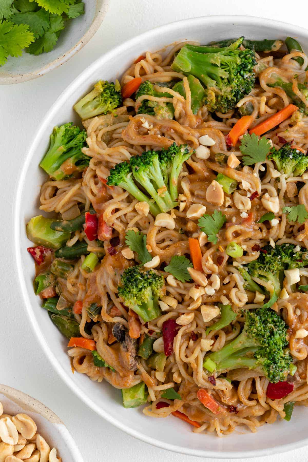 vegan peanut noodles with vegetables in a white bowl beside a bowl of cilantro and a bowl of chopped peanuts