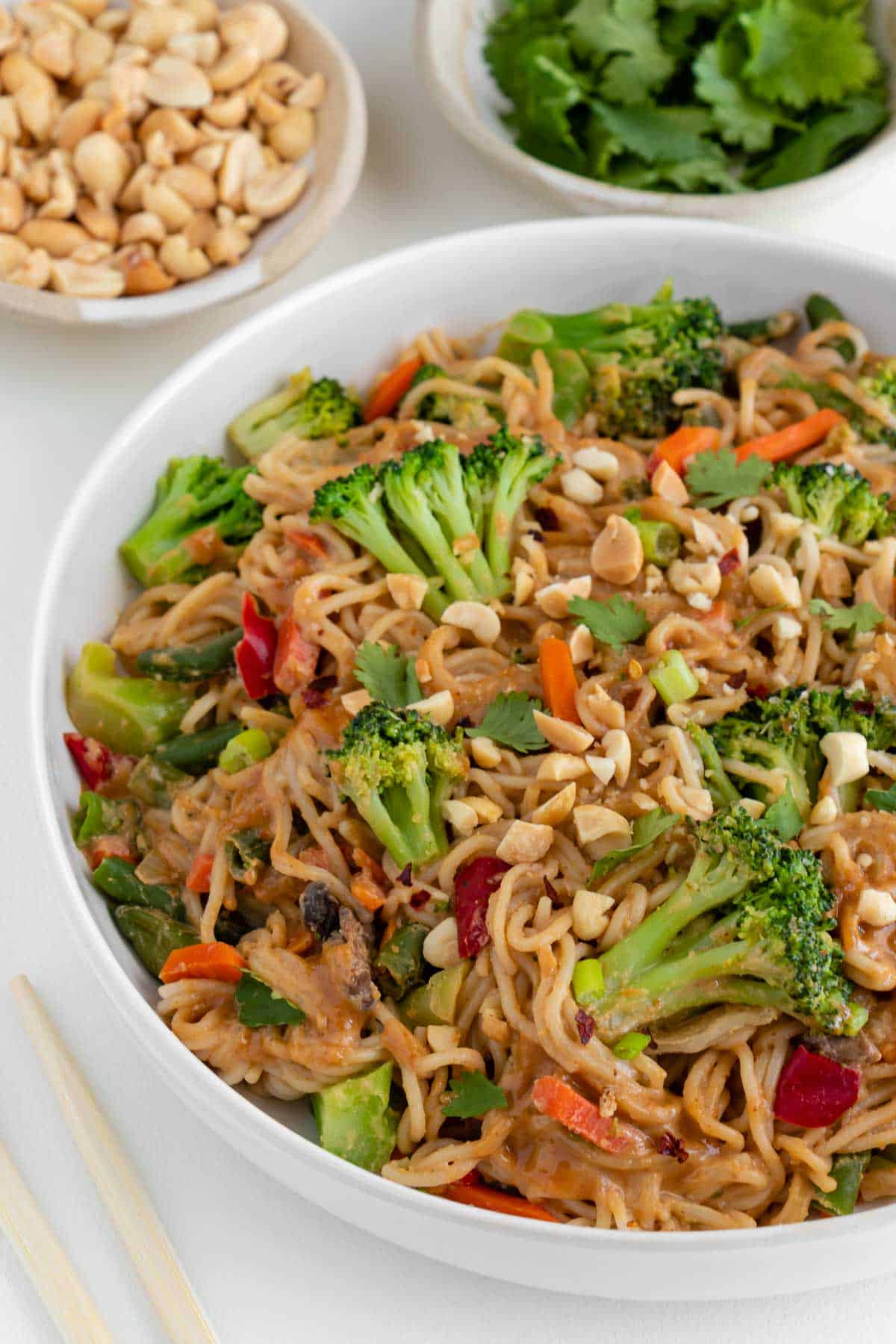 vegan peanut noodles with vegetables in a white bowl beside chopsticks, a bowl of cilantro, and a bowl of chopped peanuts