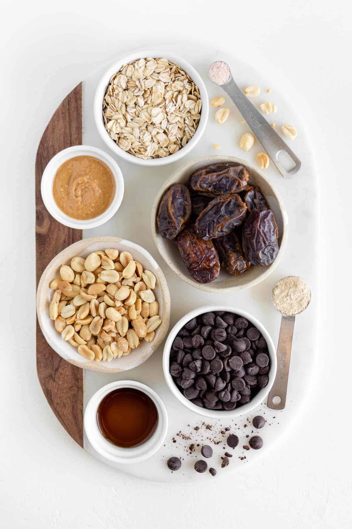 small bowls filled with oats, medjool dates, peanuts, peanut butter, chocolate chips, and vanilla extract on a marble board