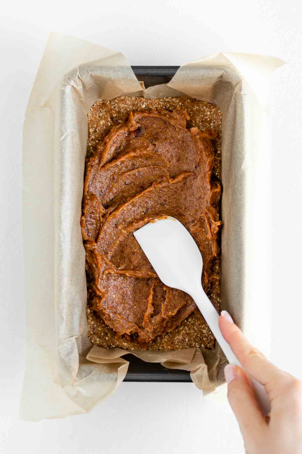 a hand holding a white spatula spreading medjool date caramel on a vegan nougat in a loaf pan