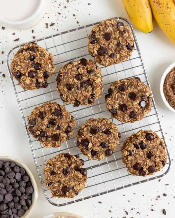 eight banana oatmeal chocolate chip cookies on a wire cooling rack surrounded by a bundle of bananas, oats, and a glass of almond milk