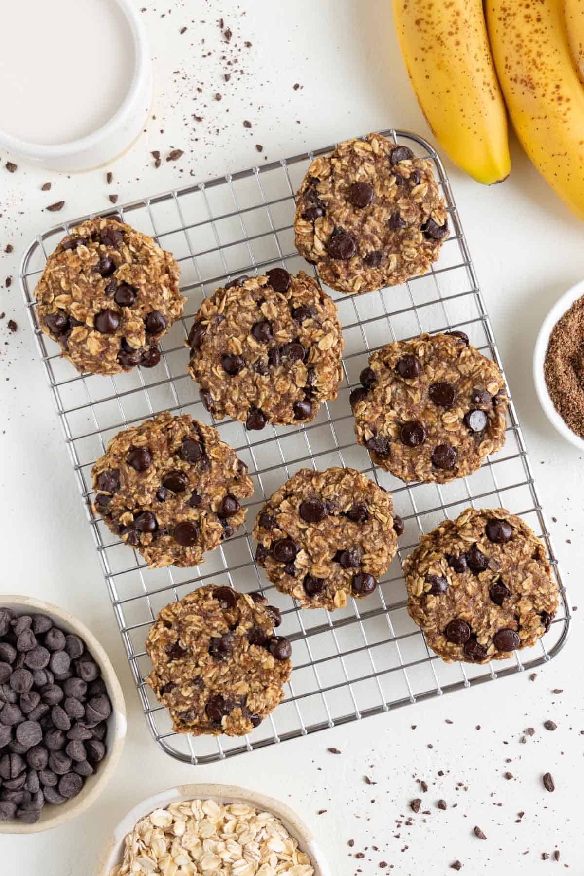 eight banana oatmeal chocolate chip cookies on a wire cooling rack surrounded by a bundle of bananas, oats, and a glass of almond milk