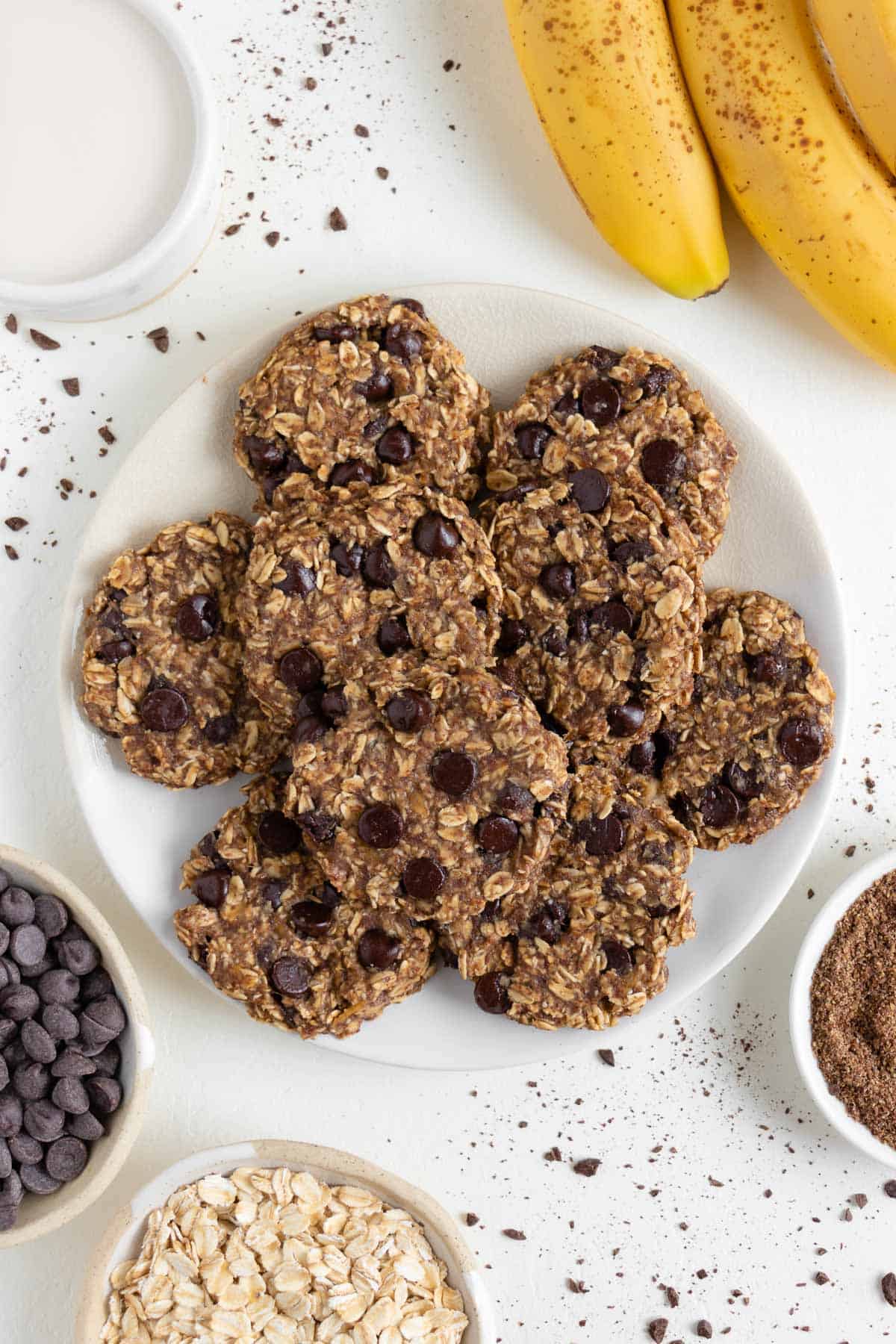 a plate of banana oatmeal chocolate chip cookies surrounded by a bundle of bananas, a glass of almond milk, a bowl of oats, and a bowl of chocolate