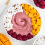 a pink raspberry mango smoothie bowl surrounded by a wooden fork and bowls of fruit