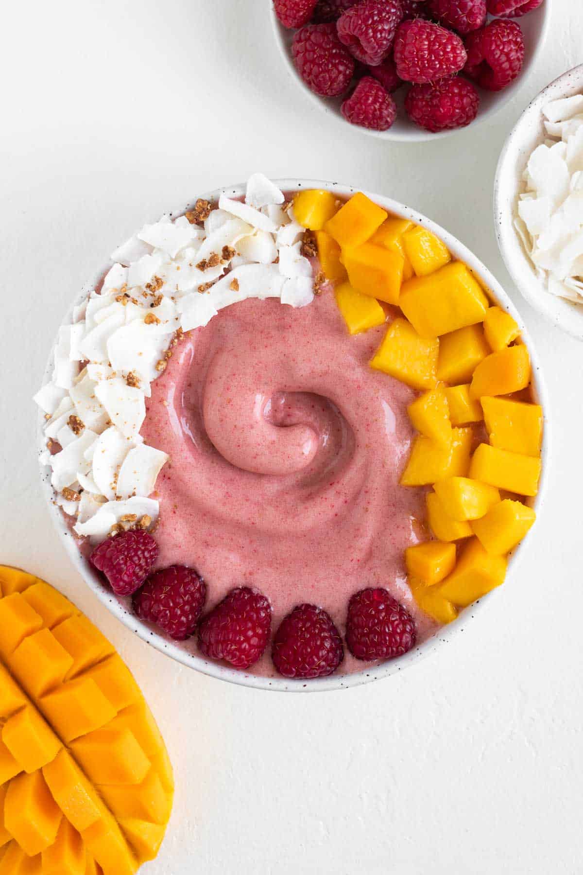 a raspberry mango smoothie bowl surrounded by a bowl of berries, a bowl of coconut, and a sliced mango