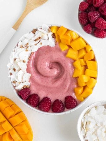 a pink raspberry mango smoothie bowl surrounded by a wooden fork and bowls of fruit
