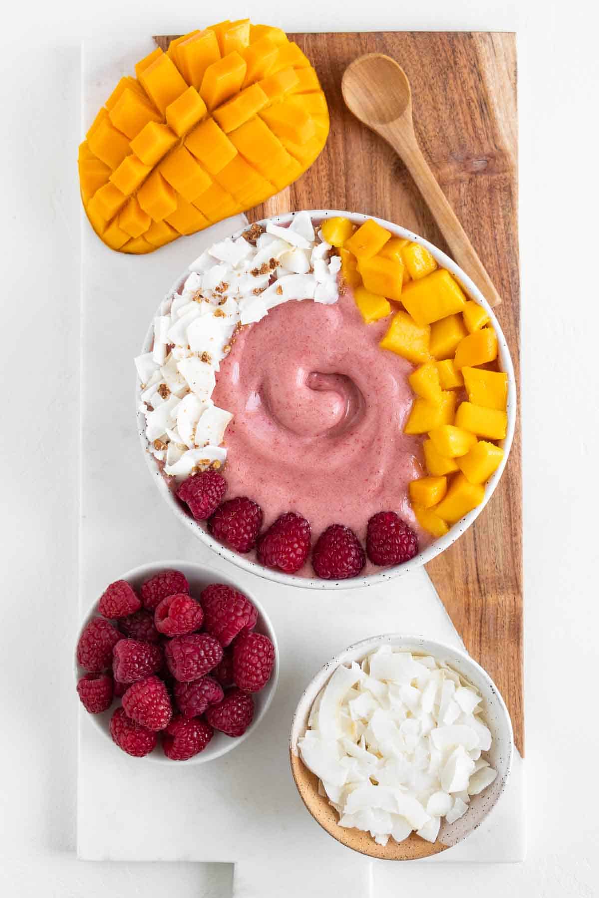 a marble and wooden cutting board topped with a raspberry mango smoothie bowl, a sliced mango, a bowl of berries, a bowl of coconut flakes, and a wooden spoon