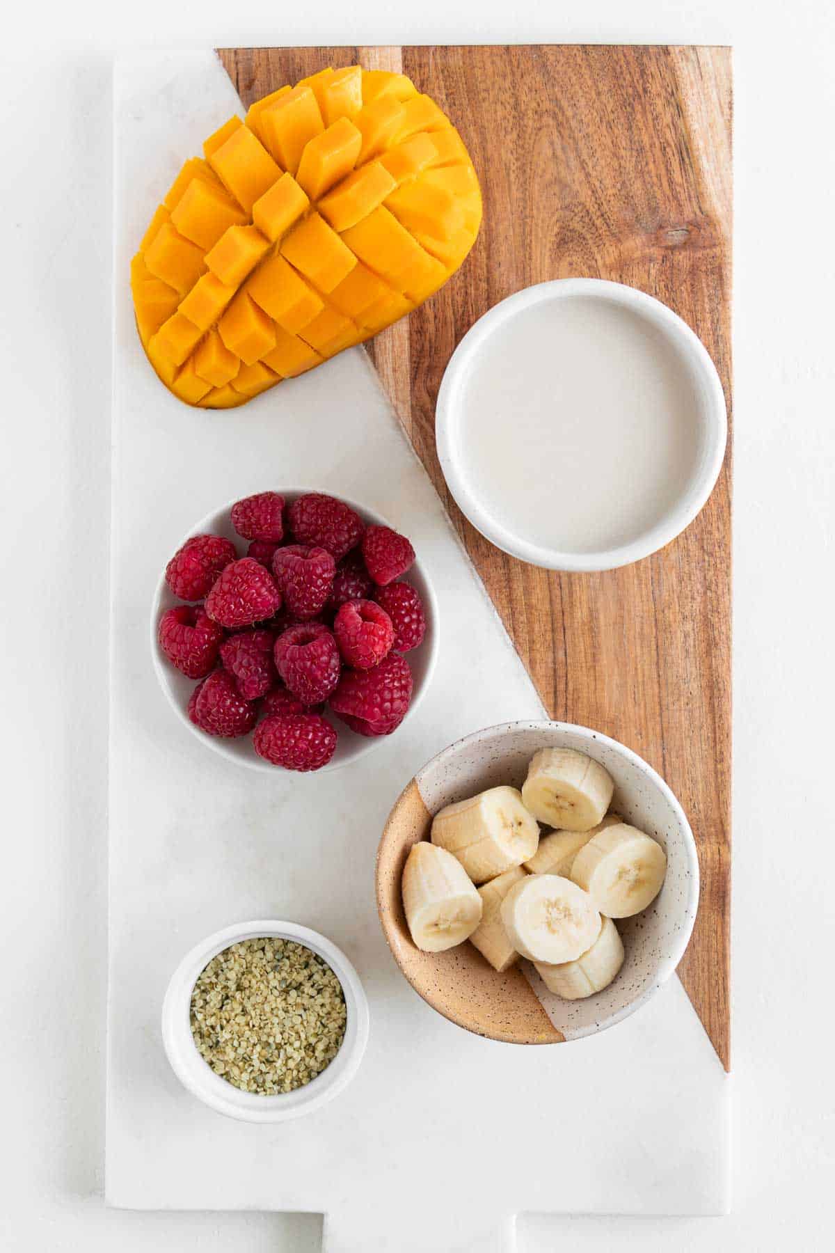 a marble and wood cutting board topped with a glass of coconut milk, bananas, berries, and hemp seeds