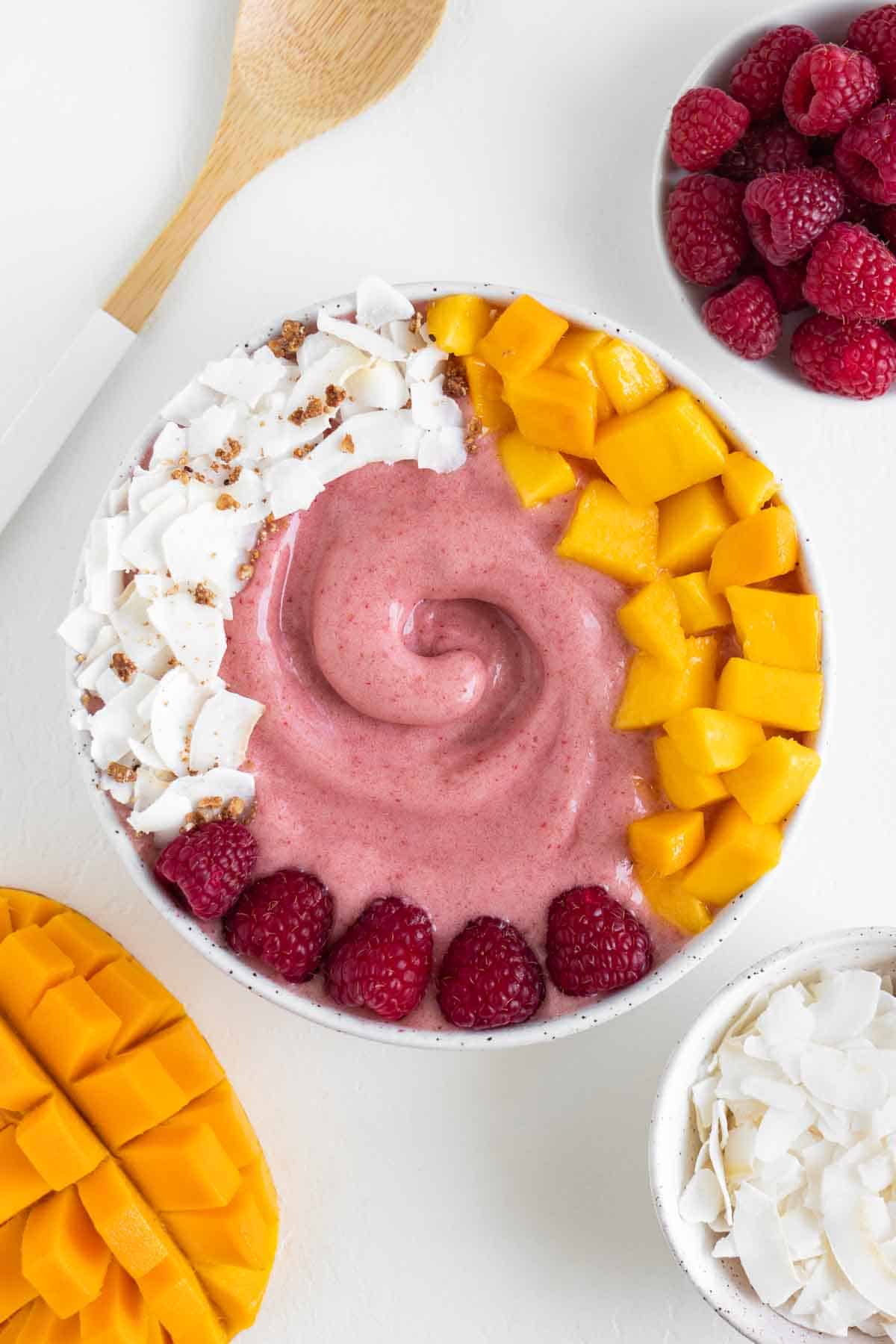 a pink raspberry mango smoothie bowl surrounded by a wooden fork and bowls of fruit