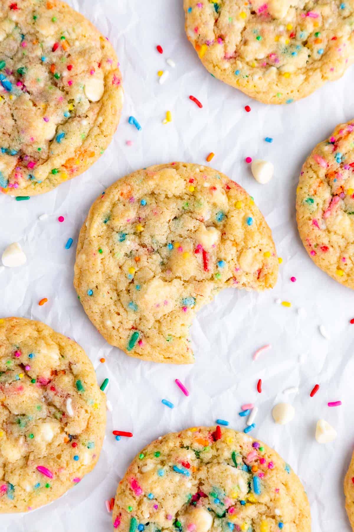 three vegan birthday cake cookies on a wire cooling rack surrounded by rainbow sprinkles and chocolate chips