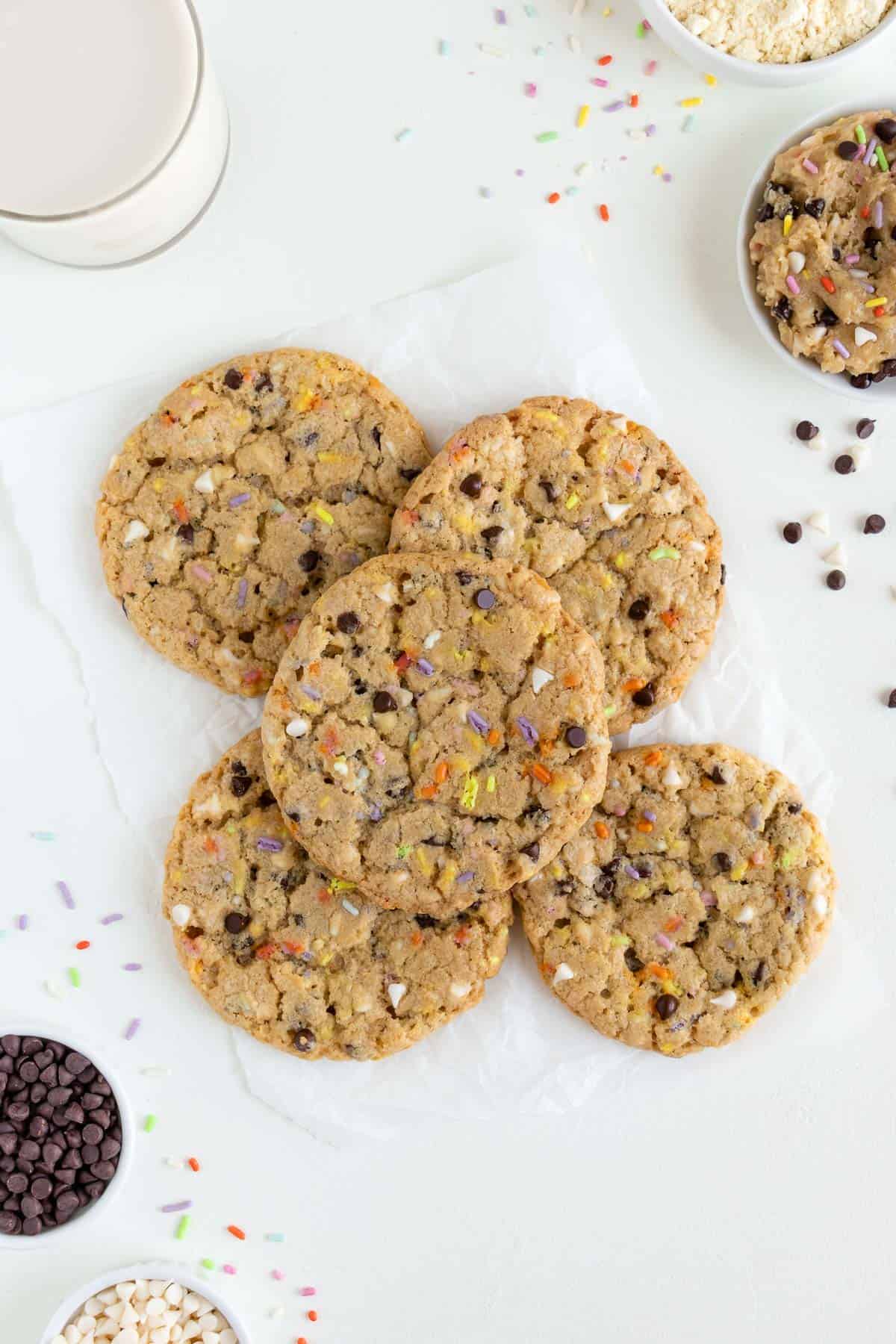 five vegan birthday cake cookies surrounded by chocolate chips, rainbow sprinkles, and white chocolate chips