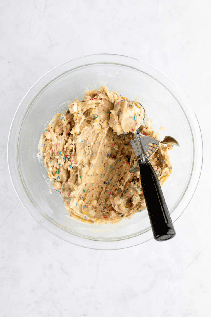 a cookie scoop inside a bowl of vegan birthday cake cookie dough with white chocolate chips and sprinkles
