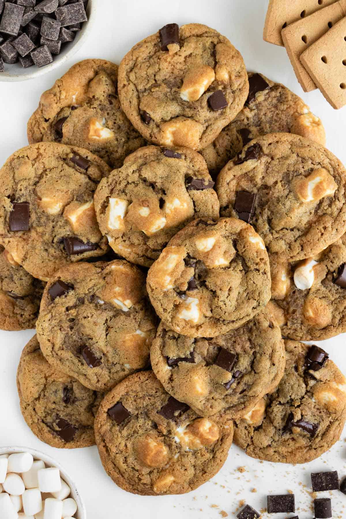 a pile of vegan s'mores cookies surrounded by a bowl of mini vegan marshmallows, graham crackers, and chocolate chunks