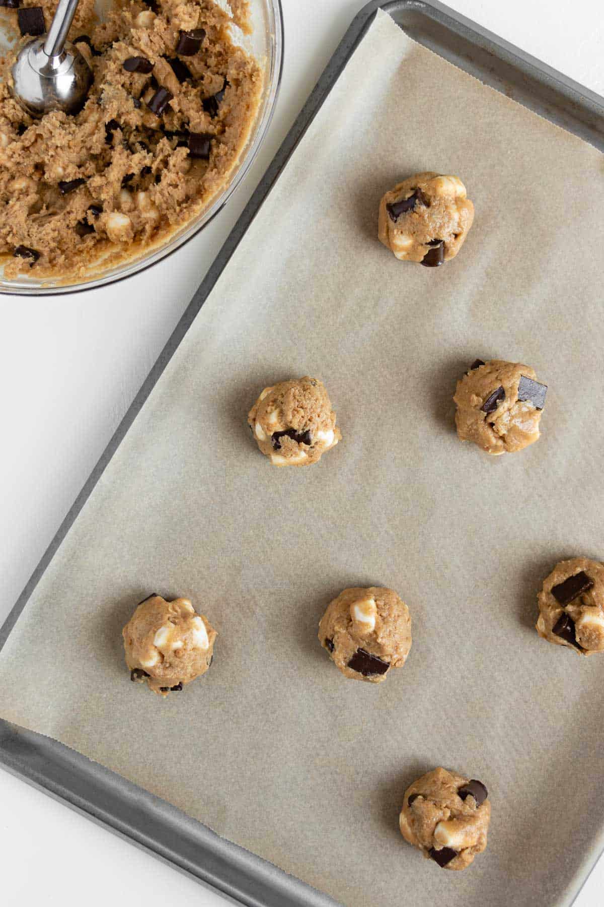 a baking sheet topped with balls of cookie dough beside a glass bowl of more cookie dough