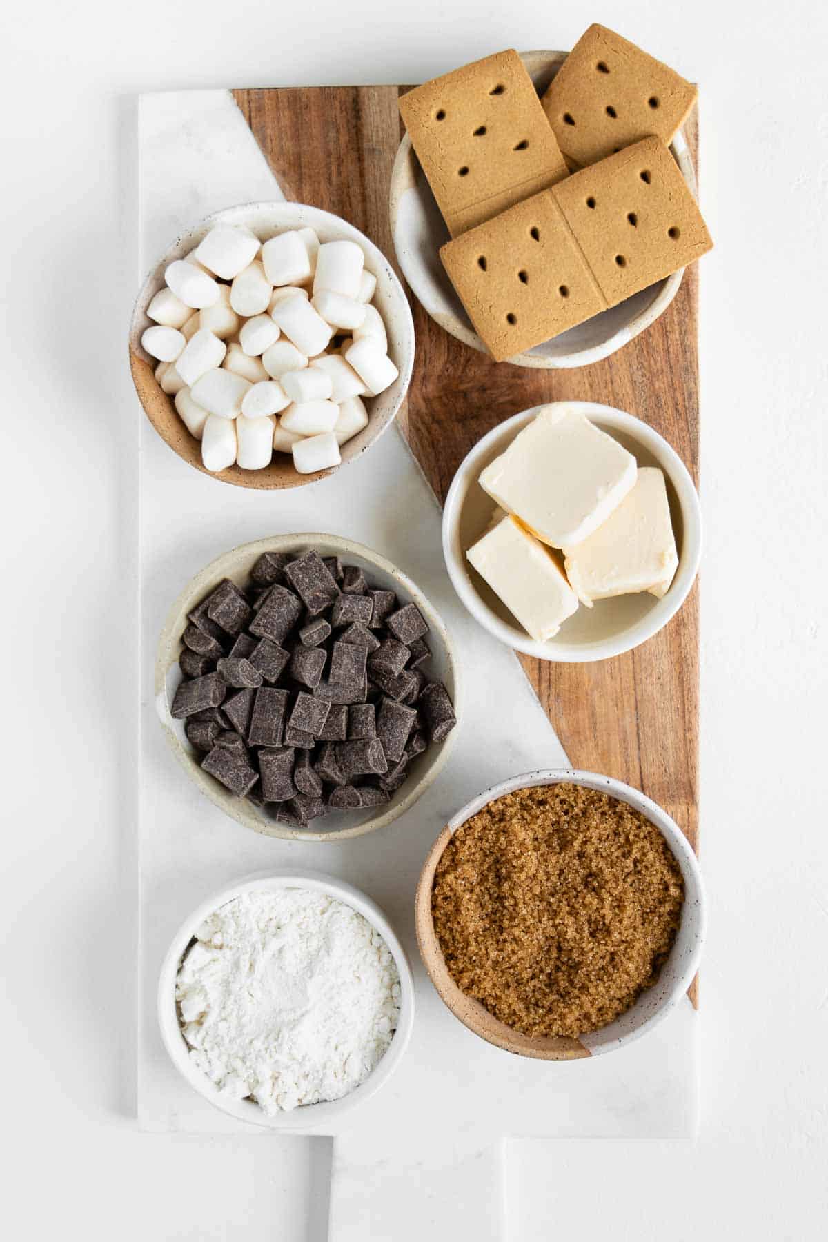 a marble and wood board topped with bowls of marshmallows, chocolate chunks, graham crackers, flour, vegan butter, and brown sugar
