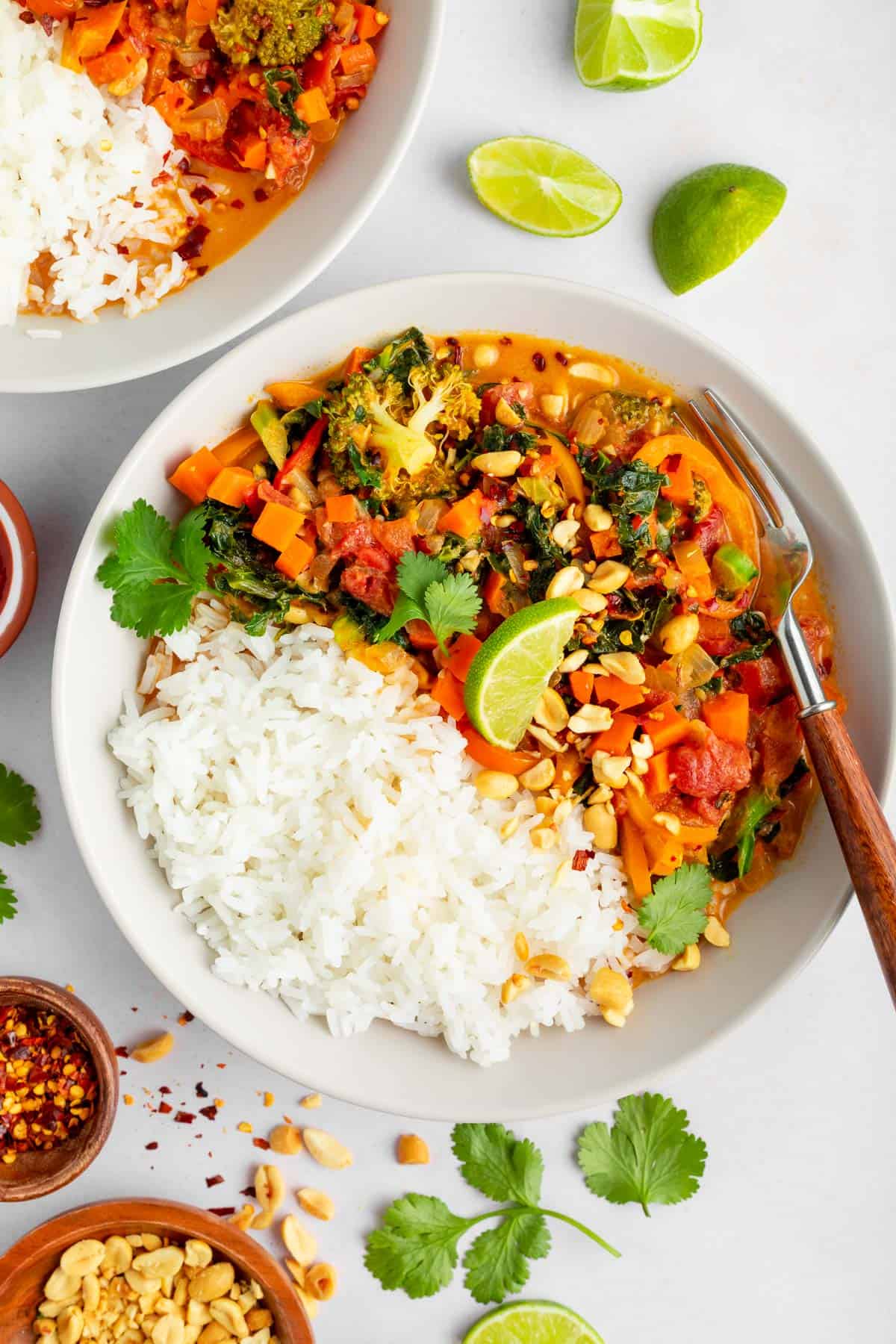 two white bowls filled with vegan thai red curry and white rice beside a bowl of cilantro