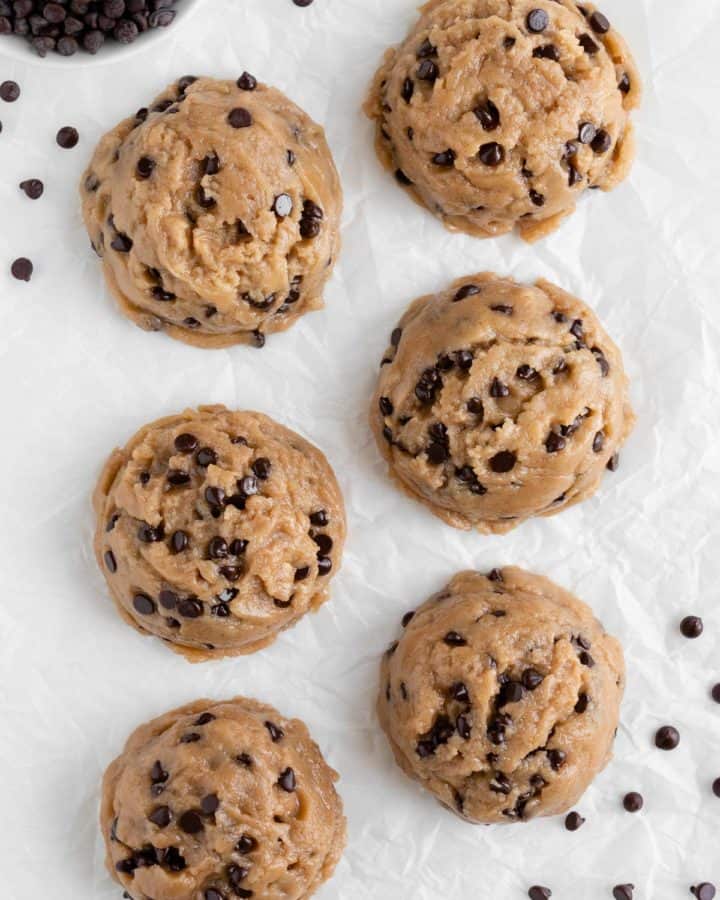 six large scoops of edible vegan cookie dough on white parchment paper surrounded by chocolate chips