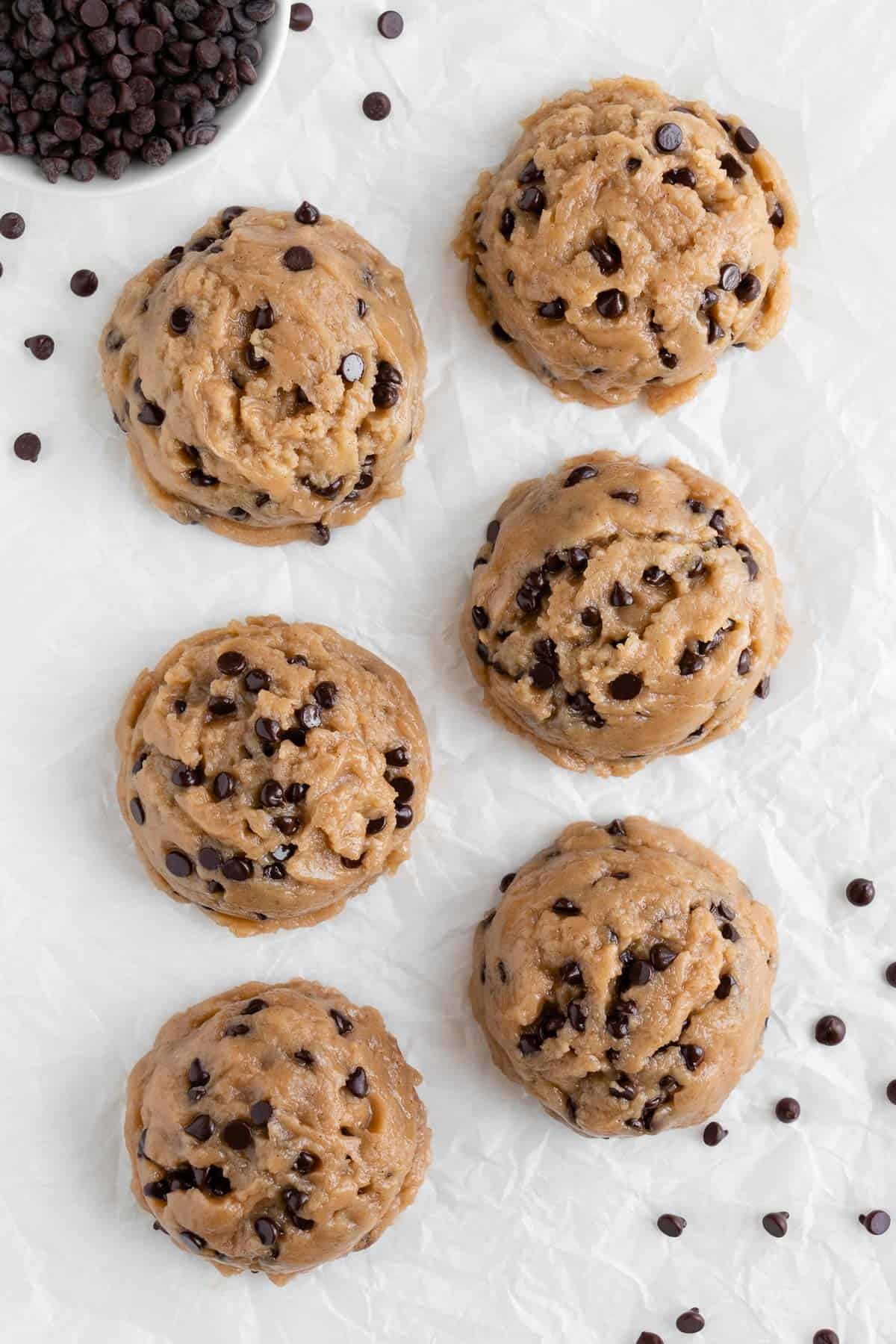 six large scoops of edible vegan cookie dough on white parchment paper surrounded by chocolate chips