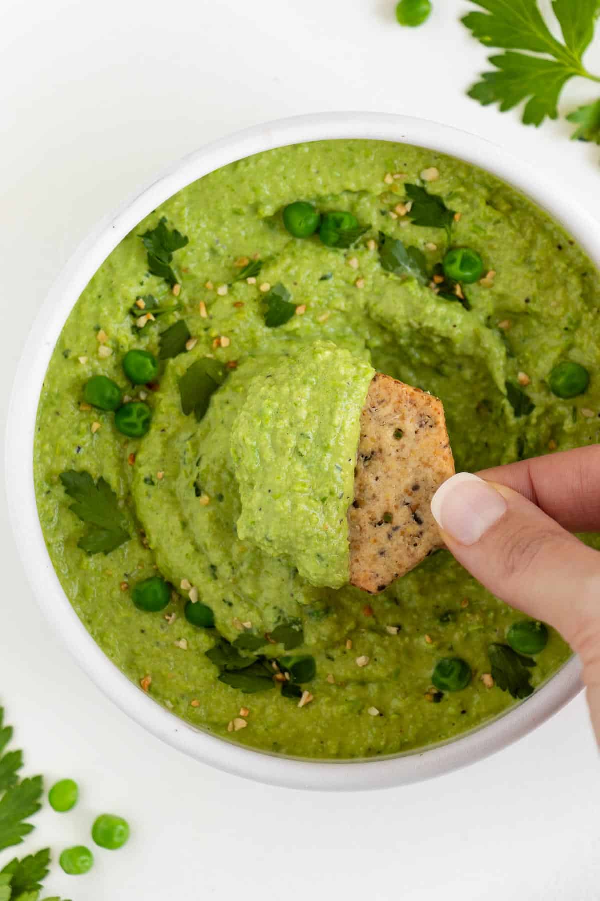 a hand scooping a cracker into a bowl of green pea dip