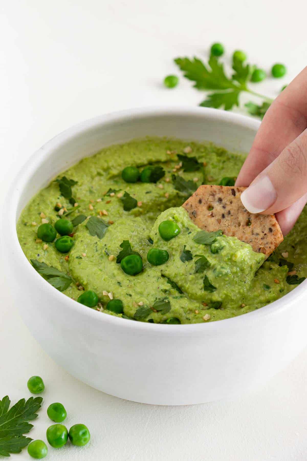 a hand scooping a cracker into a bowl of green pea dip