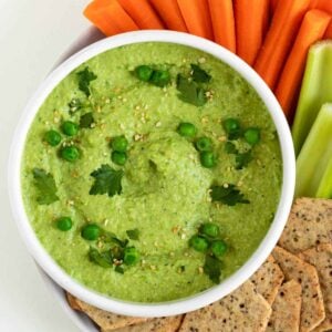 green pea hummus in a white bowl surrounded by sliced carrots, celery, and crackers