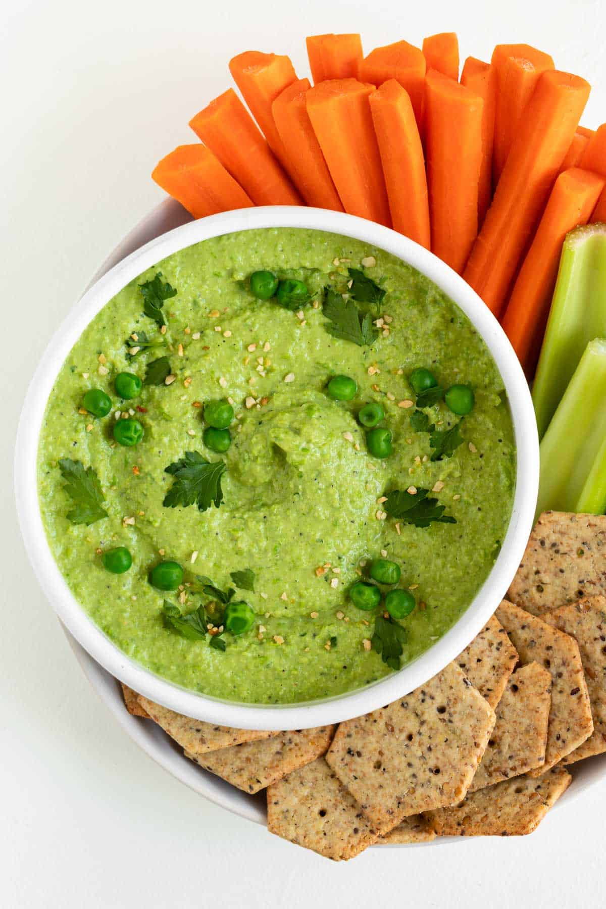 green pea hummus in a white bowl surrounded by sliced carrots, celery, and crackers