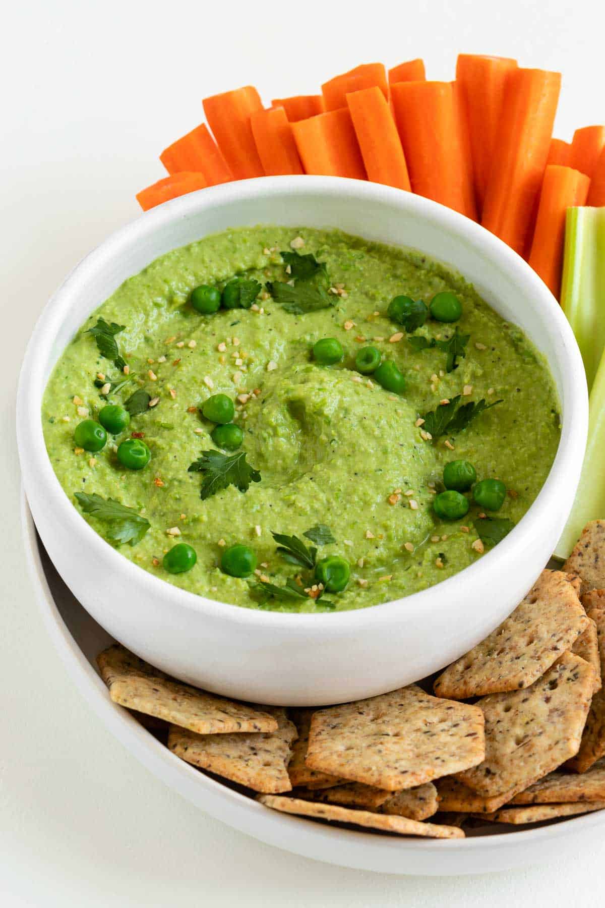 a white bowl filled with green pea hummus surrounded by sliced carrots, celery, and crackers