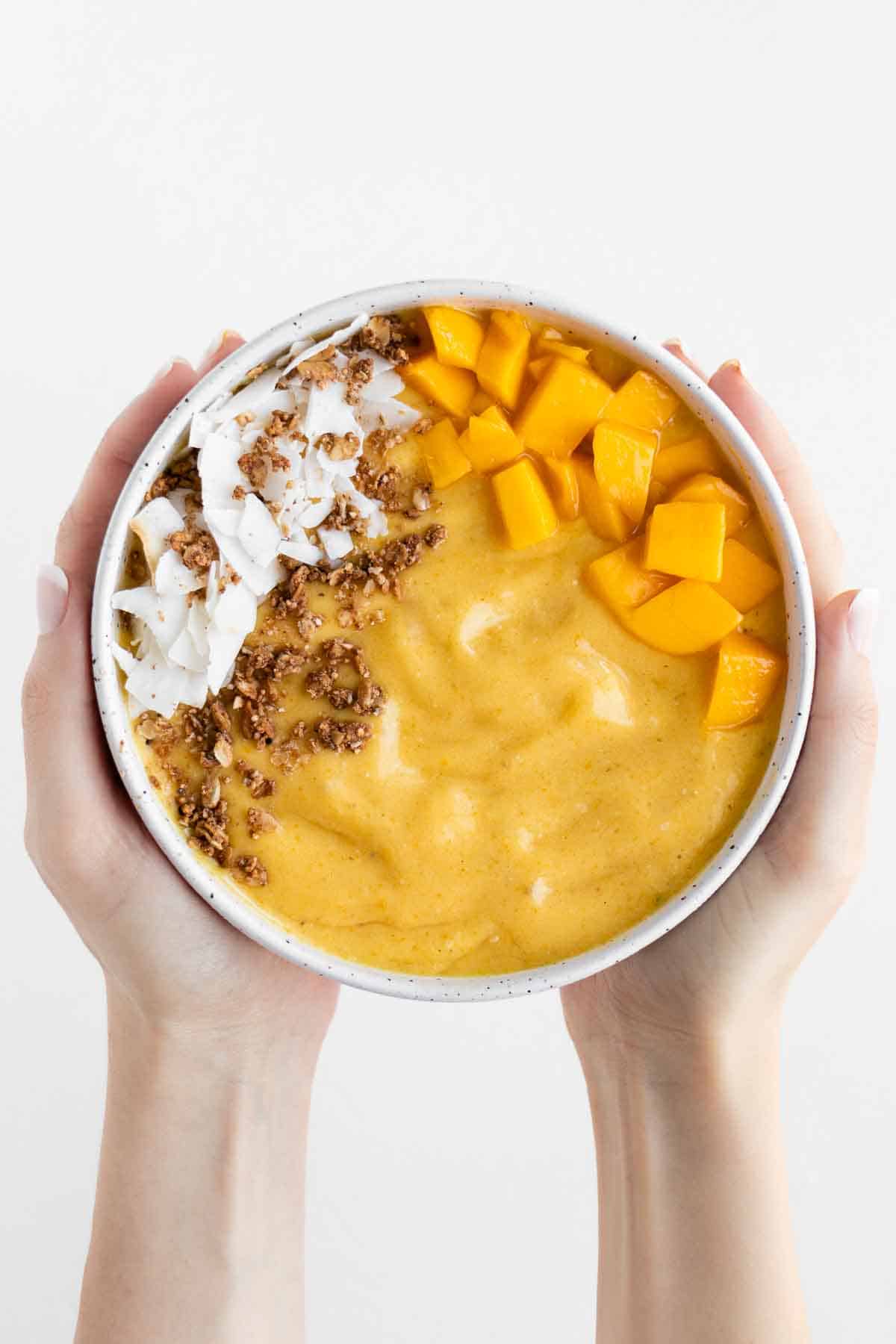 two hands holding a mango smoothie bowl topped with coconut flakes and granola