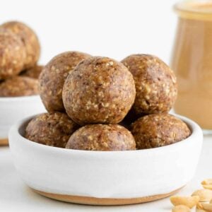 peanut butter energy balls inside a white ceramic bowl beside a jar of nut butter and chopped peanuts