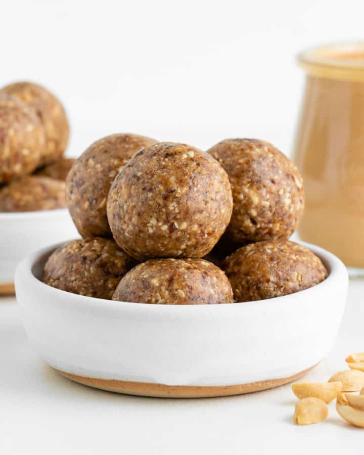 peanut butter energy balls inside a white ceramic bowl beside a jar of nut butter and chopped peanuts