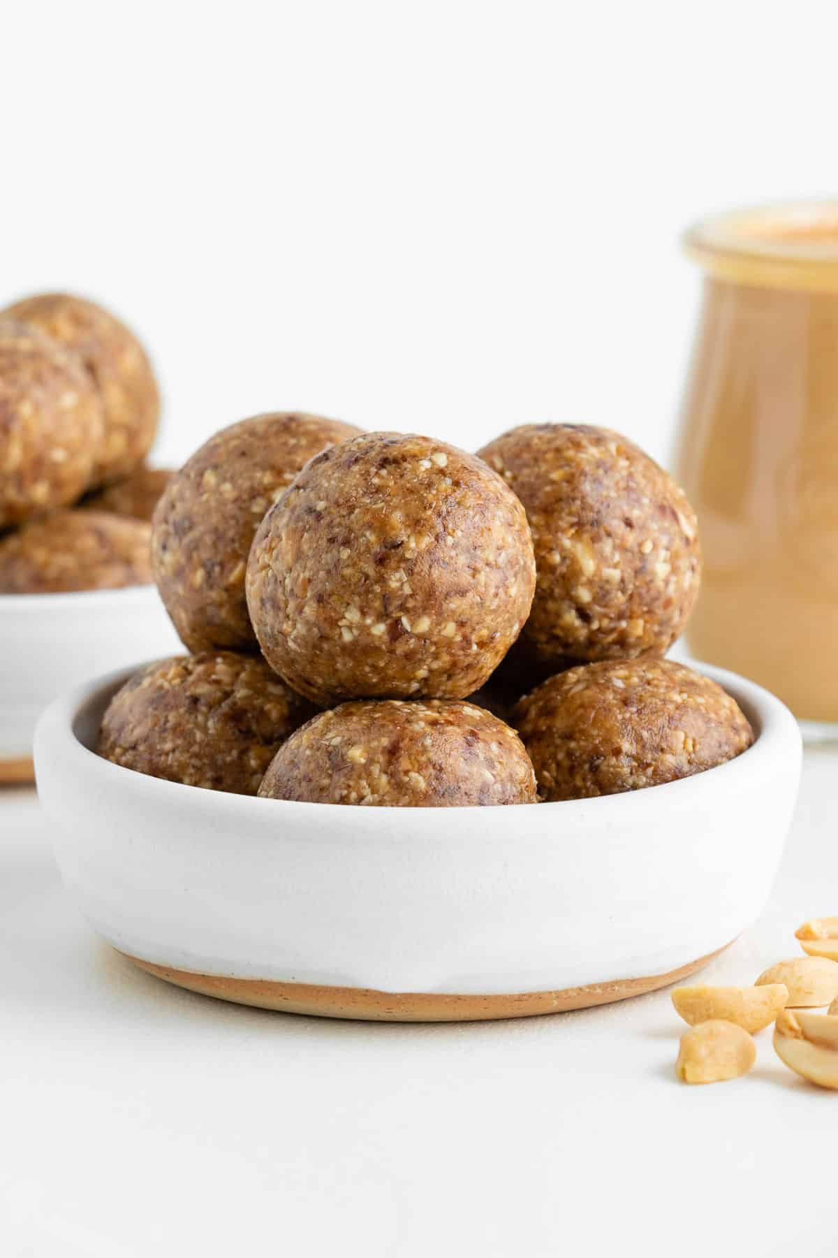 peanut butter energy balls inside a white ceramic bowl beside a jar of nut butter and chopped peanuts