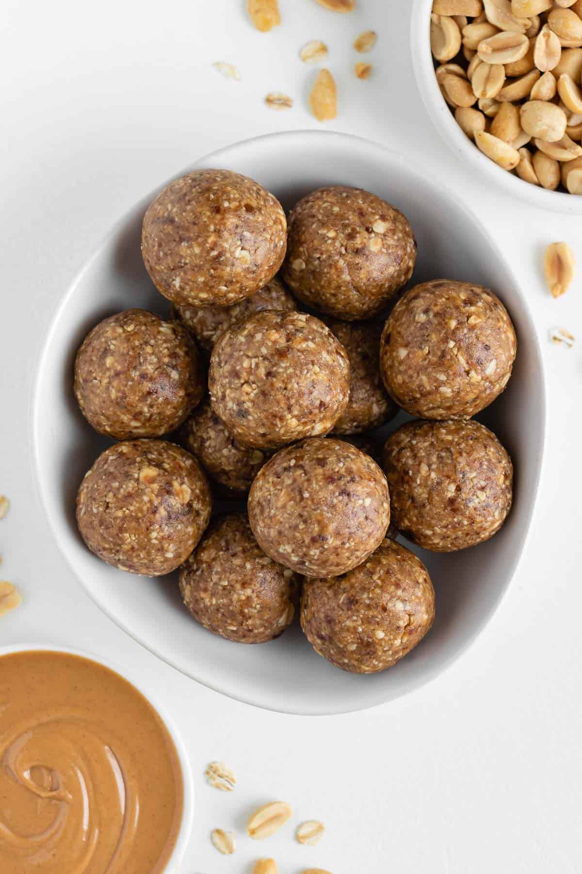 peanut butter energy balls stacked inside a white bowl surrounded by a bowl of peanut butter and a bowl of nuts