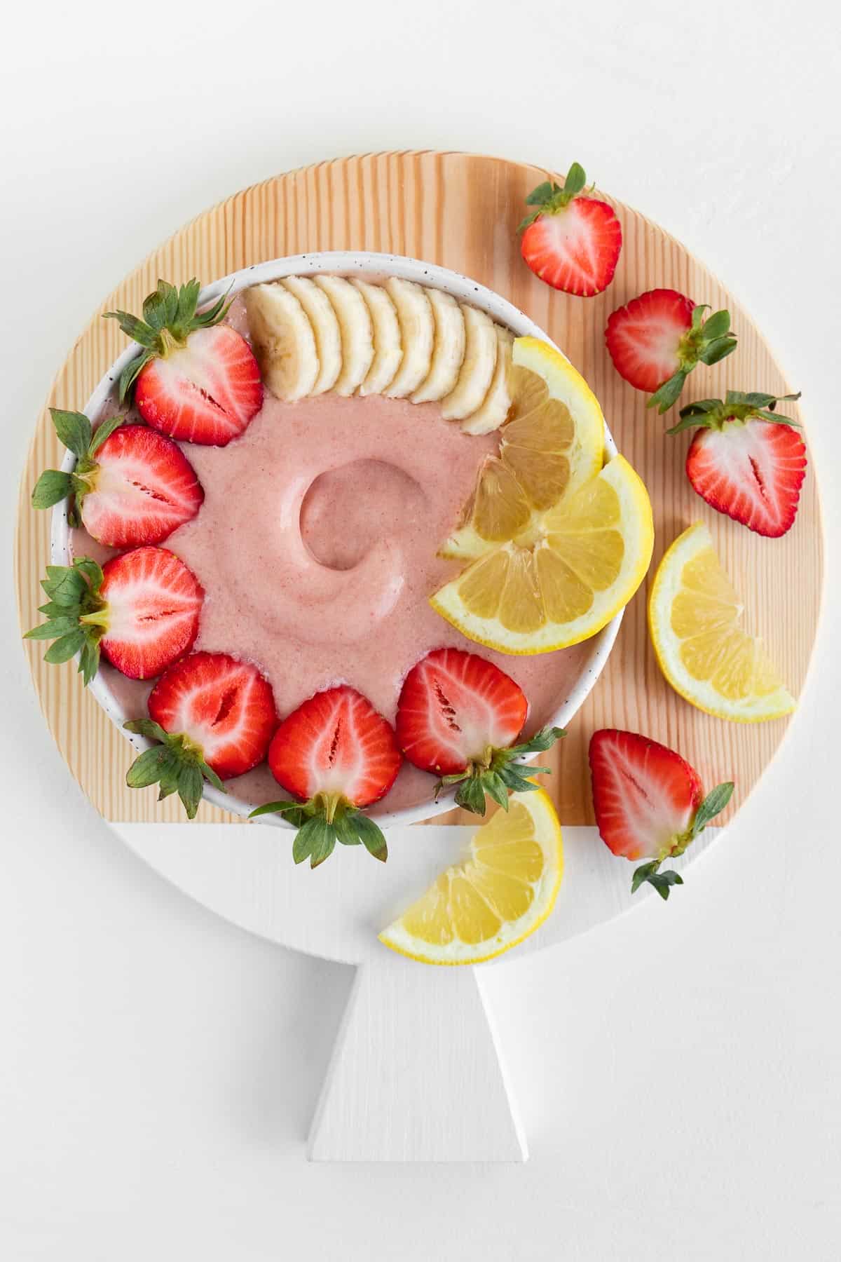 a round wooden cutting board topped with a strawberry lemonade smoothie bowl, sliced berries, and sliced lemons