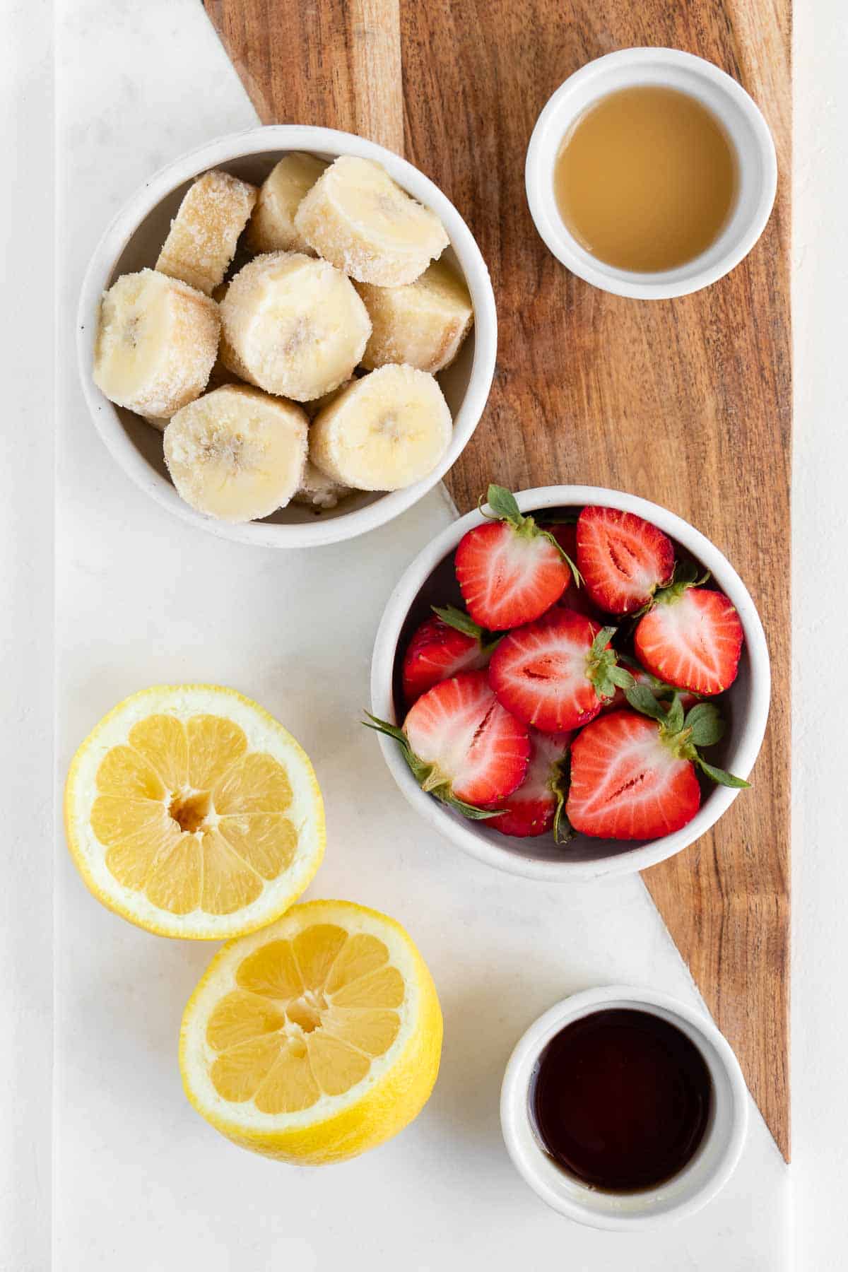 a marble and wooden board topped with a sliced lemon, a bowl of berries, a bowl of frozen bananas, a bowl of maple syrup, and a bowl of lemon juice