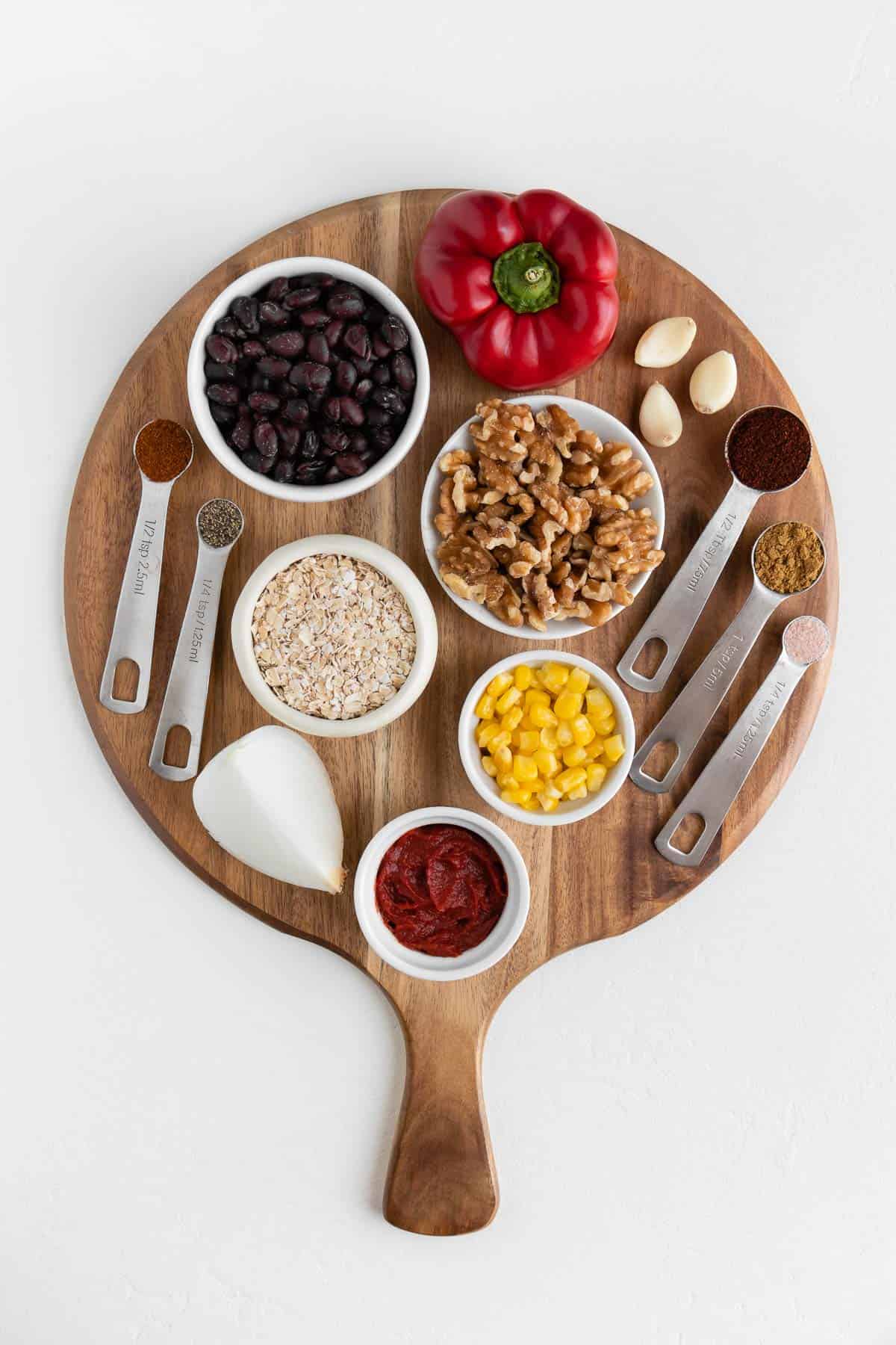 a round wooden cutting board topped with beans, corn, oats, walnuts, red bell pepper, garlic, onion, and spices