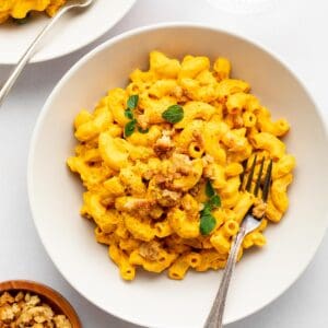 two bowls of vegan mac and cheese beside a bowl of cashews and a bowl of nutritional yeast