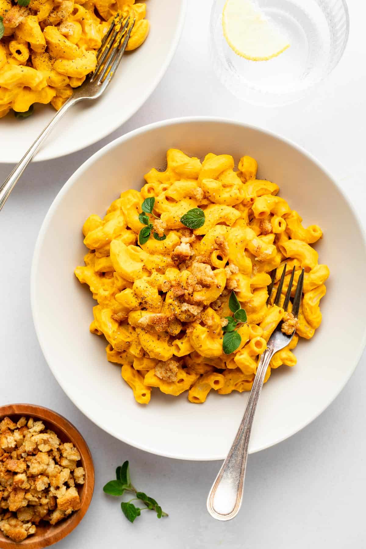 two bowls of vegan mac and cheese beside a bowl of cashews and a bowl of nutritional yeast