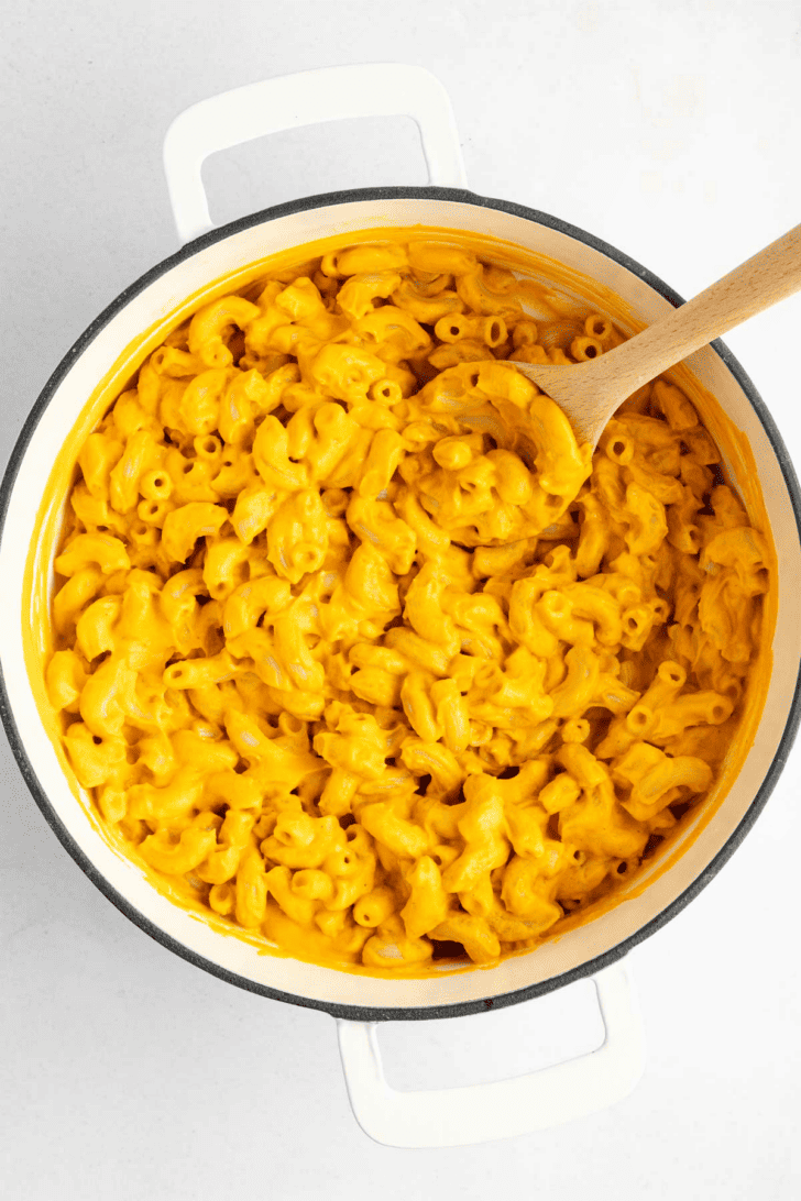 a wooden spoon mixing vegan mac and cheese inside a white le creuset dutch oven