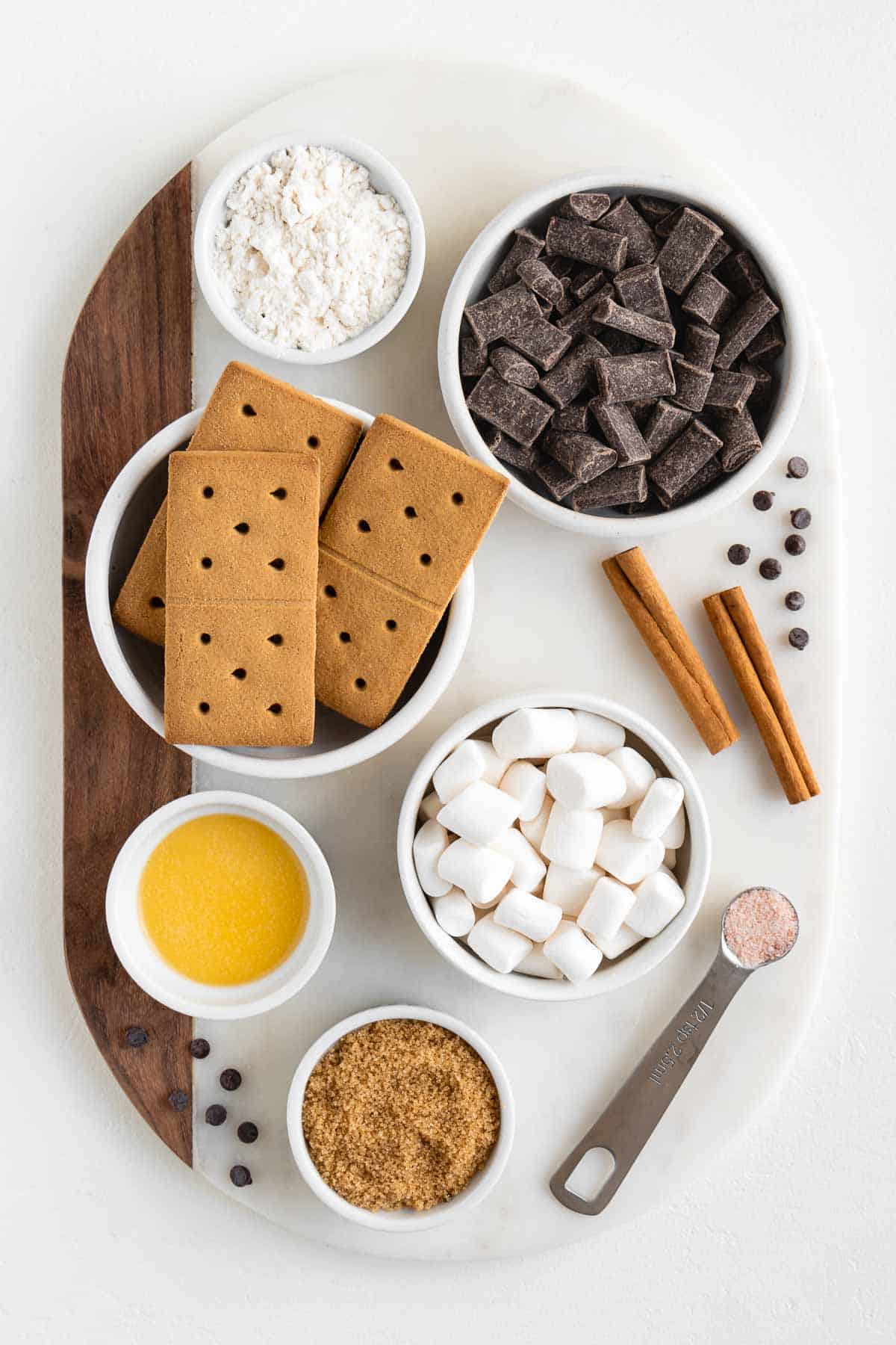 a marble cutting board topped with bowls of graham crackers, chocolate chunks, mini marshmallows, brown sugar, vegan butter, cinnamon sticks, flour, and salt