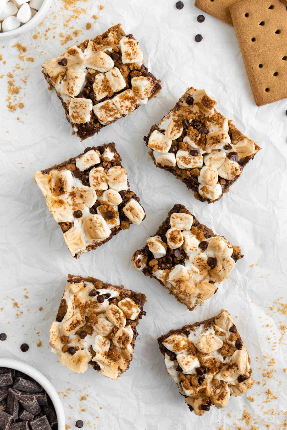 six vegan s'mores bars topped with toasted marshmallows and chocolate chips, surrounded by graham cracker crumbs and chocolate
