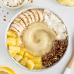 piña colada smoothie bowl beside a wooden spoon, a bowl of pineapple, a bowl of coconut flakes, and a lemon
