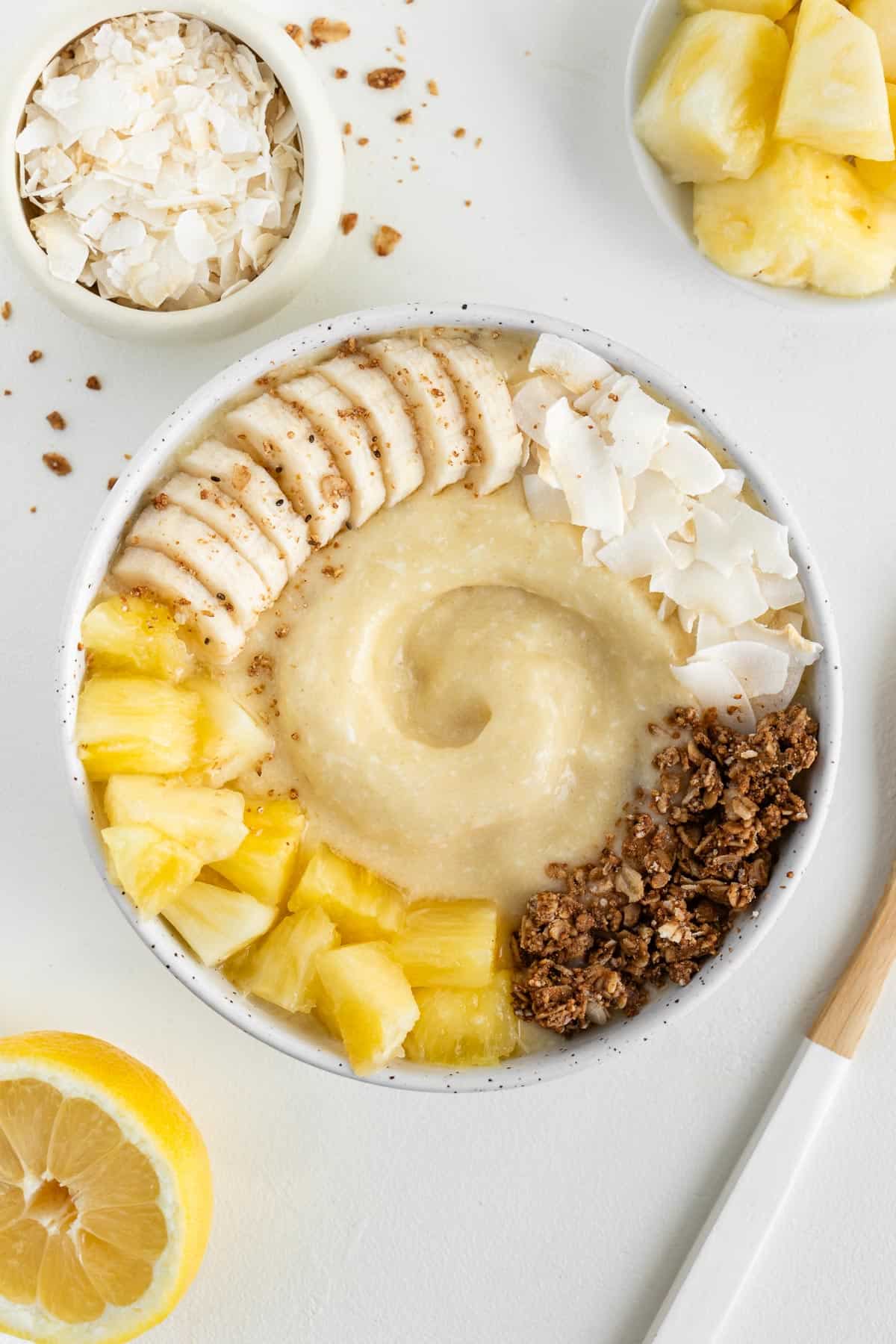 piña colada smoothie bowl beside a wooden spoon, a bowl of pineapple, a bowl of coconut flakes, and a lemon