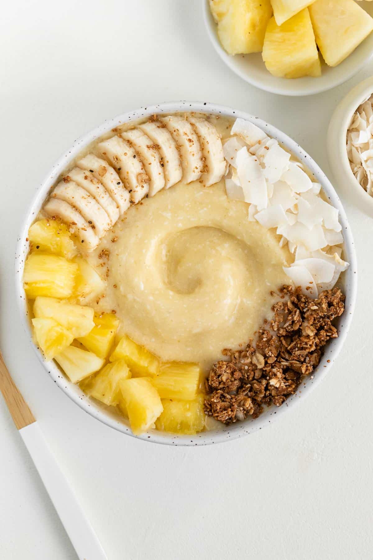 piña colada smoothie bowl beside a wooden spoon and a bowl of pineapple