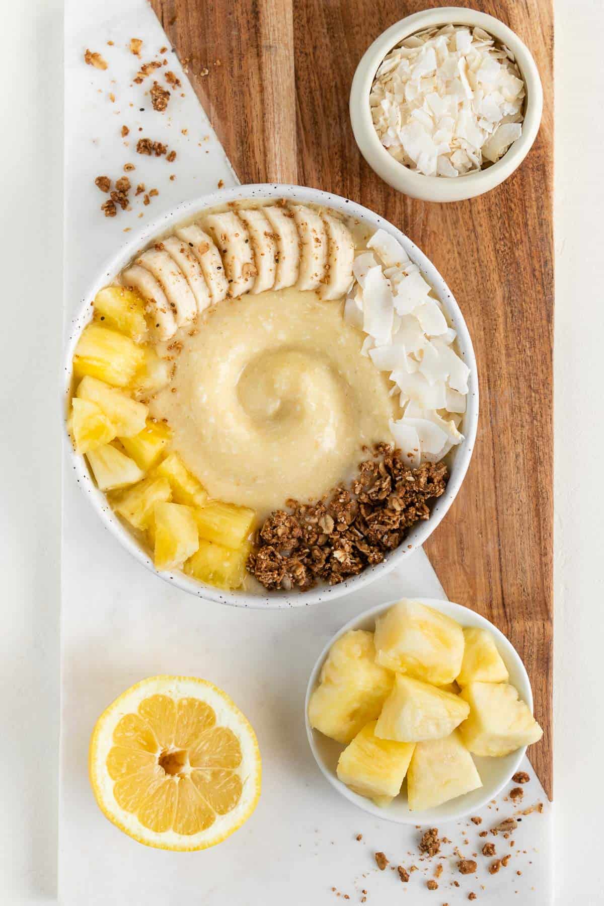 a wood and marble board topped with a piña colada smoothie bowl, bowl of coconut flakes, bowl of pineapple, and a lemon