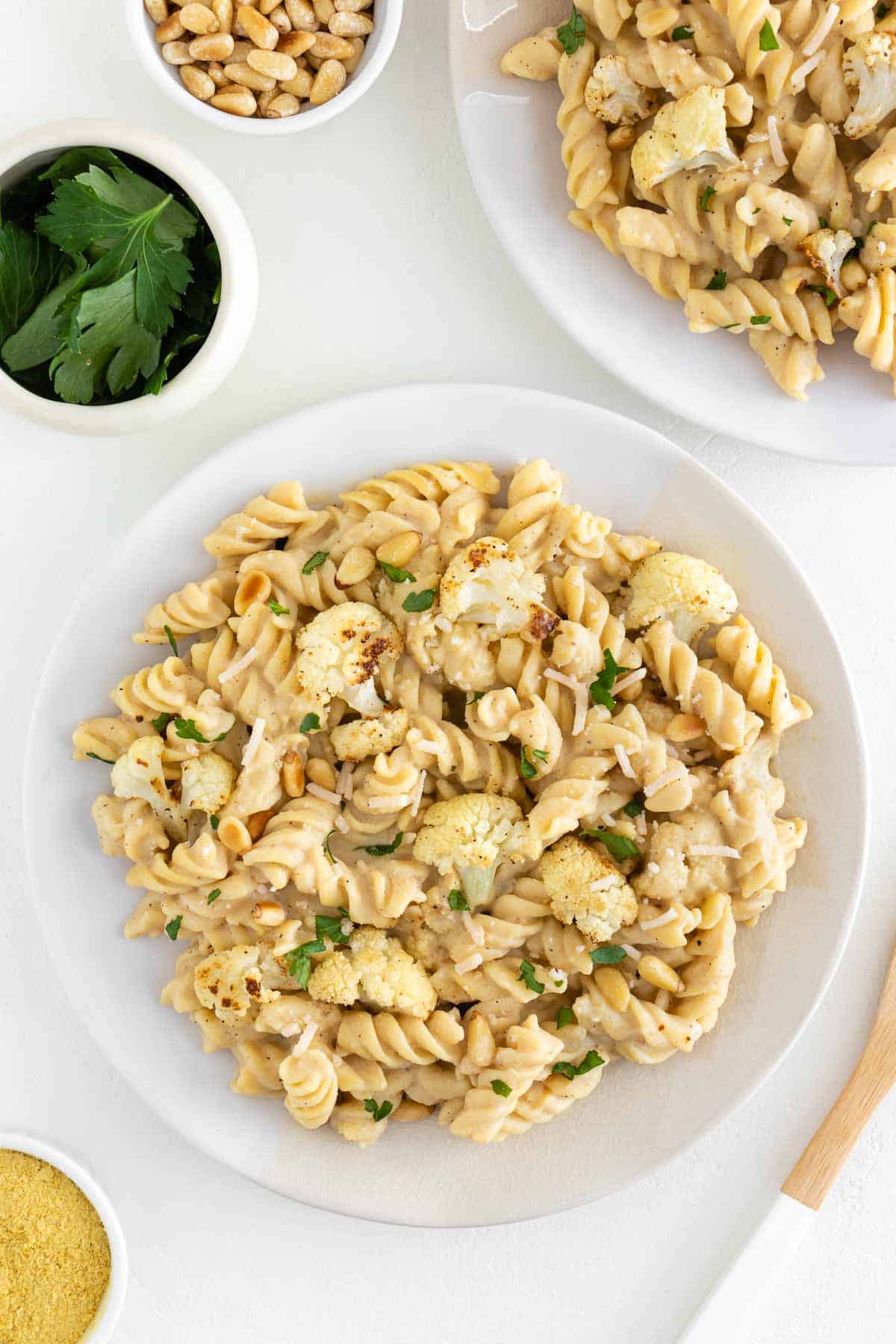 roasted cauliflower alfredo pasta on two ceramic plates beside a bowl of pine nuts and parsley